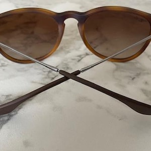 May include: Round-framed sunglasses with a tortoiseshell pattern and gradient brown lenses. The arms are silver and brown, crossed at the bottom. The glasses are set against a white and gray marble surface.