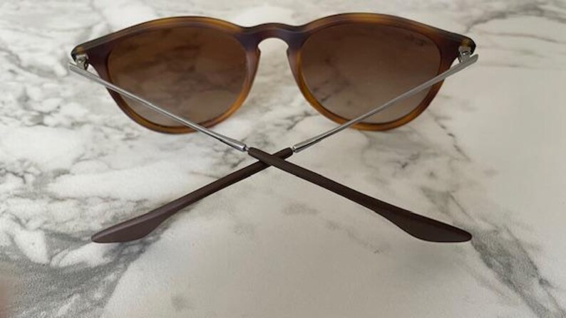 May include: Round-framed sunglasses with a tortoiseshell pattern and gradient brown lenses. The arms are silver and brown, crossed at the bottom. The glasses are set against a white and gray marble surface.