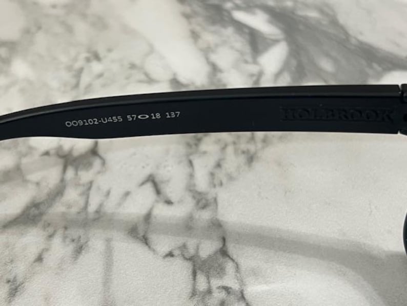 May include: Close-up of a black Holbrook sunglasses arm. The arm features the model number, lens and bridge measurements, and the brand name. The sunglasses are designed for eye protection.