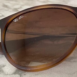 May include: Close-up of Ray-Ban sunglasses with brown frames and lenses. The sunglasses have a classic design with a rounded shape. The Ray-Ban logo is visible on the lens. The sunglasses are resting on a light-colored surface.