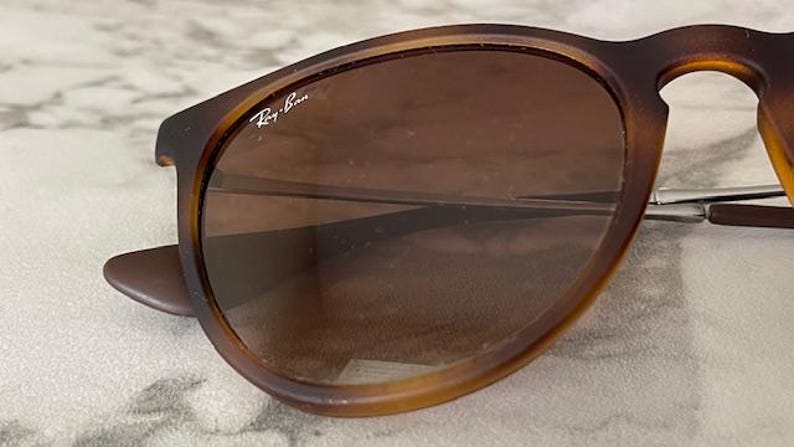 May include: Close-up of Ray-Ban sunglasses with brown frames and lenses. The sunglasses have a classic design with a rounded shape. The Ray-Ban logo is visible on the lens. The sunglasses are resting on a light-colored surface.