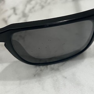 May include: Black sunglasses with dark tinted lenses. The sunglasses have a thick, rectangular frame. The lenses are slightly reflective, and the image is taken from a close-up perspective.