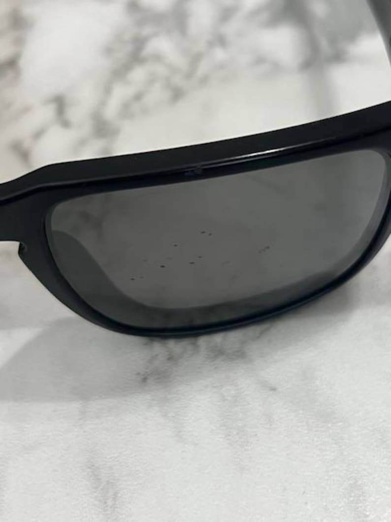 May include: Black sunglasses with dark tinted lenses. The sunglasses have a thick, rectangular frame. The lenses are slightly reflective, and the image is taken from a close-up perspective.