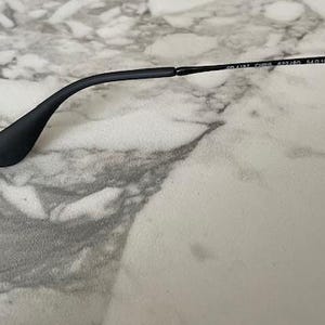 May include: Black sunglasses with a matte finish. The temple arm curves gracefully, ending in a rounded tip. The text on the arm reads "RB 4187 CHINA 54/18 140 3N". The frame rests on a marble-patterned surface.