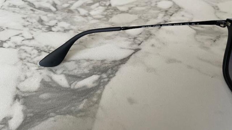 May include: Black sunglasses with a matte finish. The temple arm curves gracefully, ending in a rounded tip. The text on the arm reads "RB 4187 CHINA 54/18 140 3N". The frame rests on a marble-patterned surface.