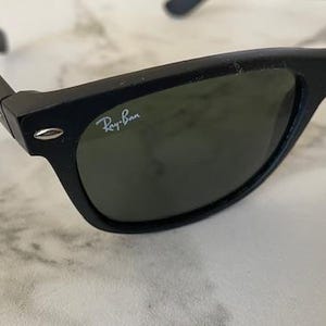 May include: Black sunglasses with green lenses. The Ray-Ban logo is visible on the right lens. The sunglasses are on a white and gray marble surface.