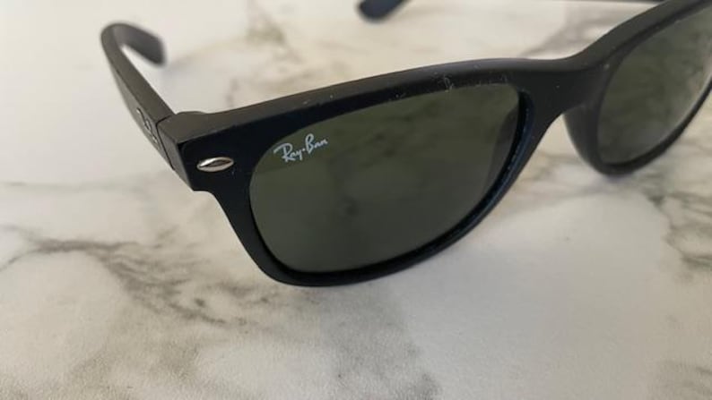 May include: Black sunglasses with green lenses. The Ray-Ban logo is visible on the right lens. The sunglasses are on a white and gray marble surface.