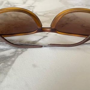 May include: Brown sunglasses with a tortoiseshell frame and brown lenses. The sunglasses have a classic design with a curved browline and temples. The arms are a darker brown color and connect to the lenses with a silver hinge.