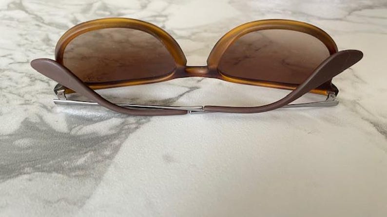 May include: Brown sunglasses with a tortoiseshell frame and brown lenses. The sunglasses have a classic design with a curved browline and temples. The arms are a darker brown color and connect to the lenses with a silver hinge.