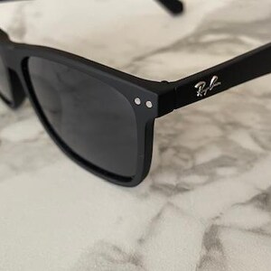 Pu&ograve; includere: Occhiali da sole neri con lenti scure. Gli occhiali hanno una finitura opaca e una montatura rettangolare. Il logo Ray-Ban &egrave; visibile sull'asta. Gli occhiali poggiano su una superficie di marmo bianco e grigio.