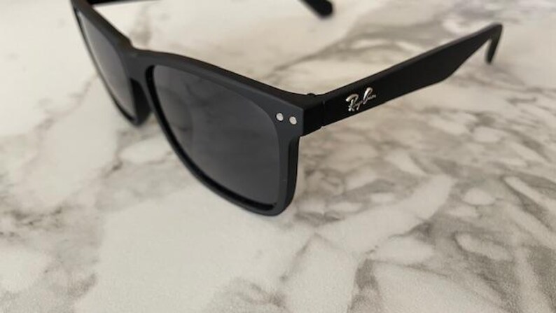 Pu&ograve; includere: Occhiali da sole neri con lenti scure. Gli occhiali hanno una finitura opaca e una montatura rettangolare. Il logo Ray-Ban &egrave; visibile sull'asta. Gli occhiali poggiano su una superficie di marmo bianco e grigio.