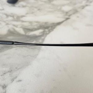 May include: Black sunglasses with dark lenses. The frame is matte black with a slender arm that curves slightly. The Ray-Ban logo is visible on the arm. The sunglasses are resting on a light-colored, marble-like surface.
