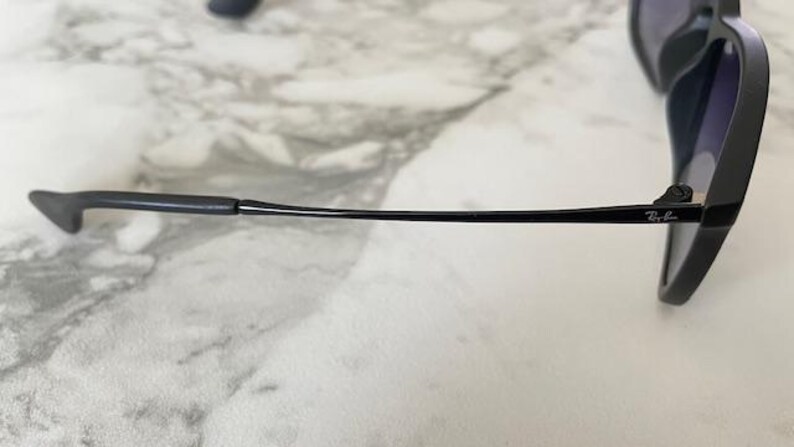 May include: Black sunglasses with dark lenses. The frame is matte black with a slender arm that curves slightly. The Ray-Ban logo is visible on the arm. The sunglasses are resting on a light-colored, marble-like surface.