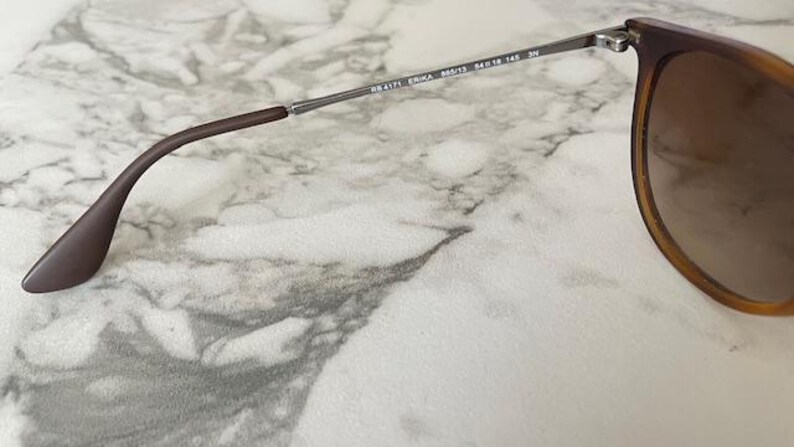 May include: Close-up of Ray-Ban Erika sunglasses. The sunglasses have a brown frame and gradient lenses. The temples are a dark brown color and connect to the lenses with silver hinges. The model number is visible on the frame.