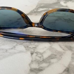 May include: Tortoiseshell-patterned sunglasses with blue-tinted lenses. The frames have a dark brown and amber pattern. The arms are black and feature the brand name. The sunglasses are resting on a marble surface.