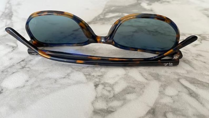 May include: Tortoiseshell-patterned sunglasses with blue-tinted lenses. The frames have a dark brown and amber pattern. The arms are black and feature the brand name. The sunglasses are resting on a marble surface.