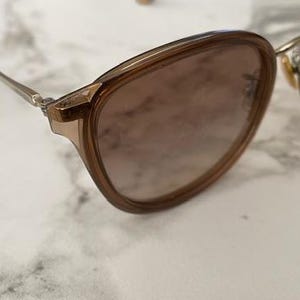 May include: A pair of sunglasses with a brown frame and lenses. The glasses have a rounded shape and thin, silver-colored arms. The lenses have a gradient tint, transitioning from a darker shade at the top to a lighter shade at the bottom.