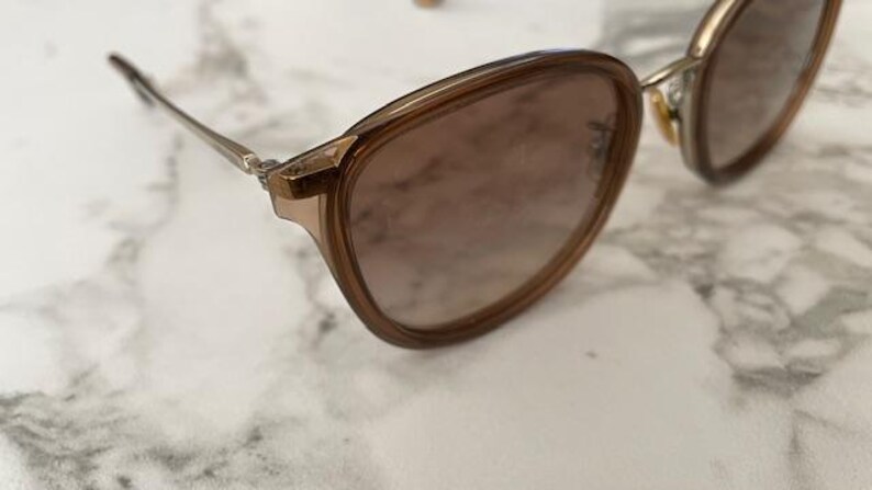 May include: A pair of sunglasses with a brown frame and lenses. The glasses have a rounded shape and thin, silver-colored arms. The lenses have a gradient tint, transitioning from a darker shade at the top to a lighter shade at the bottom.