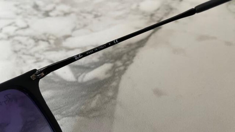 May include: Close-up of black sunglasses with purple lenses. The frame arm is visible, displaying the Ray-Ban logo and "MADE IN ITALY" text. The sunglasses are set against a light-colored, marbled background.