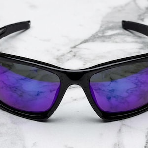 May include: Black sunglasses with purple and blue reflective lenses. The sunglasses have a rectangular frame and a glossy finish. The lenses reflect the surrounding environment, creating a vibrant color effect. The sunglasses are designed for eye protection.