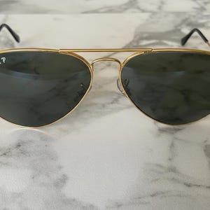 May include: Gold-framed aviator sunglasses with dark green lenses. The Ray-Ban logo is visible on the left lens. The sunglasses have a double bridge and black earpieces. The lenses are teardrop-shaped.