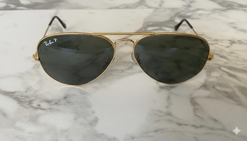 May include: Gold-framed aviator sunglasses with dark green lenses. The Ray-Ban logo is visible on the left lens. The sunglasses have a double bridge and black earpieces. The lenses are teardrop-shaped.