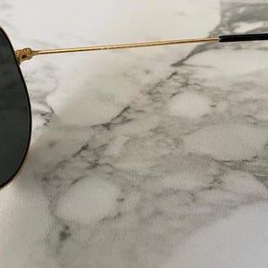 May include: Gold-rimmed sunglasses with dark lenses. The sunglasses have a classic aviator shape with a thin gold frame and black earpieces. The lenses are a dark gray color, and the sunglasses are set against a marble background.