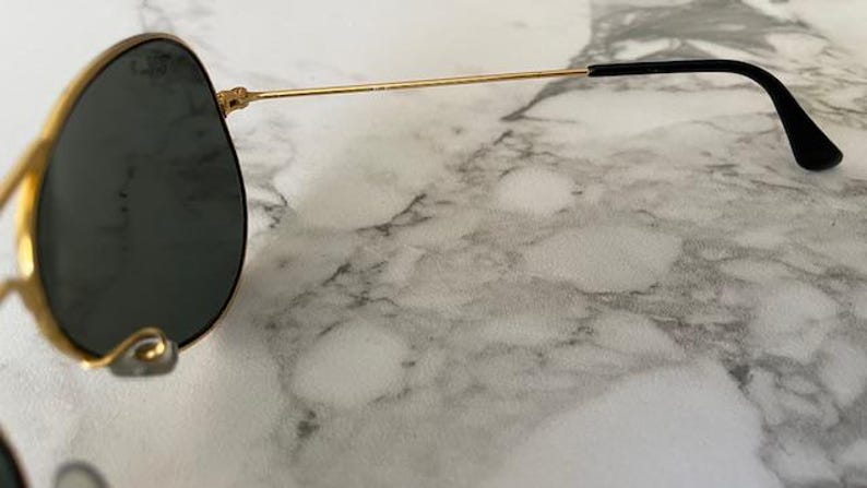 May include: Gold-rimmed sunglasses with dark lenses. The sunglasses have a classic aviator shape with a thin gold frame and black earpieces. The lenses are a dark gray color, and the sunglasses are set against a marble background.