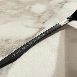 May include: Close-up of black sunglasses with gray lenses. The arm of the glasses displays the model number RB 4179 601-S/82 62▢13 140 3P.