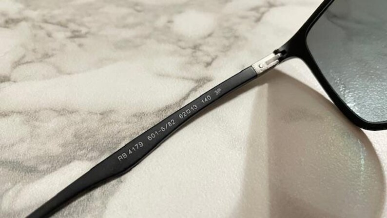 May include: Close-up of black sunglasses with gray lenses. The arm of the glasses displays the model number RB 4179 601-S/82 62▢13 140 3P.