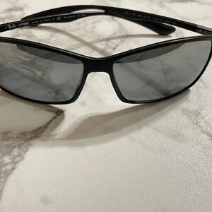May include: Black sunglasses with a sleek, modern design. The frames are black, and the lenses are a reflective silver color. The sunglasses are resting on a white and gray marble surface.