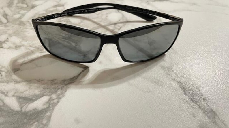 May include: Black sunglasses with a sleek, modern design. The frames are black, and the lenses are a reflective silver color. The sunglasses are resting on a white and gray marble surface.