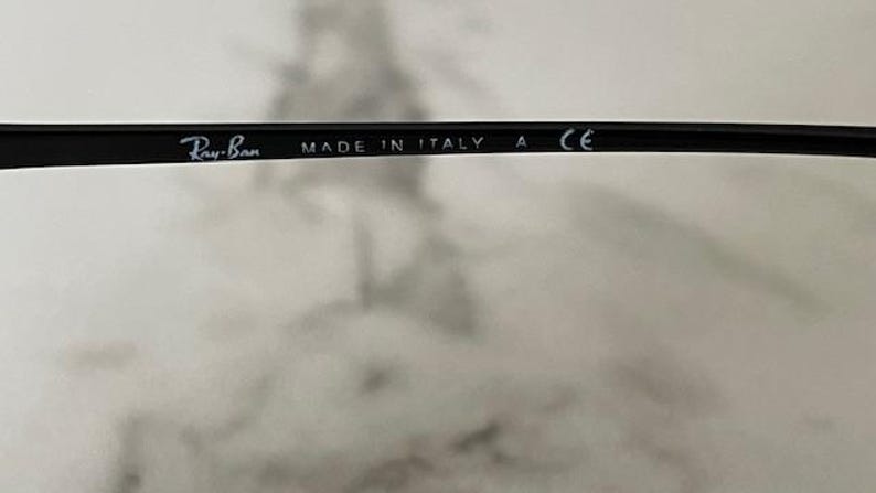 Puede incluir: Montura de gafas negra con el logotipo de Ray-Ban y el texto "MADE IN ITALY" y "CE". La montura es elegante y moderna, adecuada para lentes graduadas o gafas de sol.