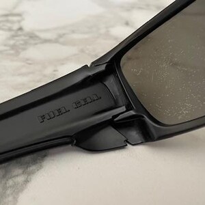 May include: Close-up of black sunglasses with a rectangular frame. The text "FUEL CELL" is embossed on the temple. The lenses are a dark grey colour.