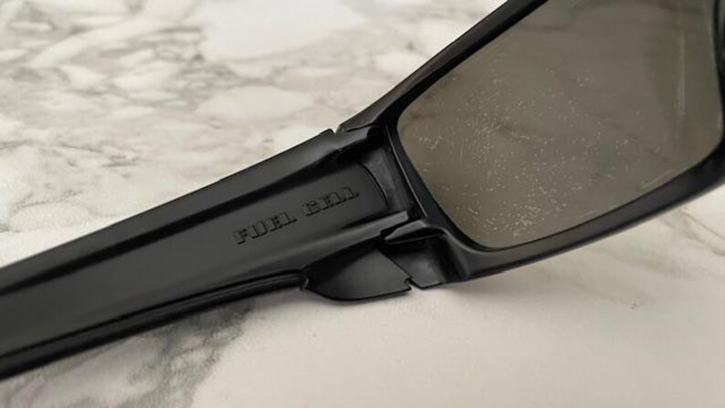 May include: Close-up of black sunglasses with a rectangular frame. The text "FUEL CELL" is embossed on the temple. The lenses are a dark grey colour.