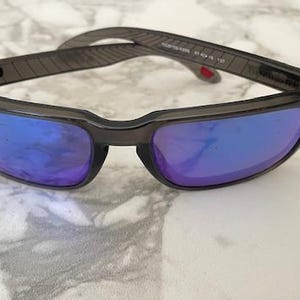 May include: A pair of sunglasses with a dark gray frame and blue-purple lenses. The temples have a textured design. The sunglasses are resting on a white and gray marble surface.