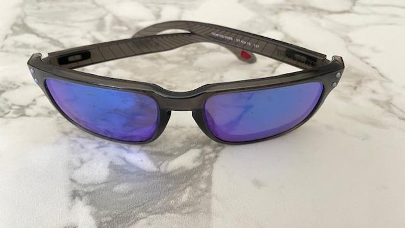 May include: A pair of sunglasses with a dark gray frame and blue-purple lenses. The temples have a textured design. The sunglasses are resting on a white and gray marble surface.