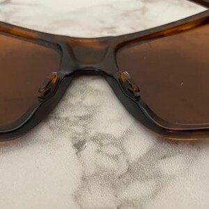 Op de afbeelding: Close-up van een zonnebril met een bruin schildpadmontuur en bruine glazen. De zonnebril heeft een rechthoekige vorm en een donkere brug. De neuspads zijn zichtbaar. De afbeelding is vanuit een lage hoek genomen.