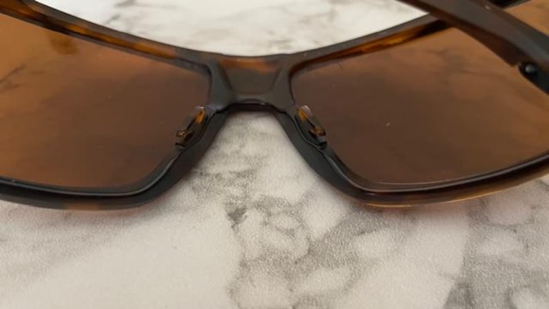 Op de afbeelding: Close-up van een zonnebril met een bruin schildpadmontuur en bruine glazen. De zonnebril heeft een rechthoekige vorm en een donkere brug. De neuspads zijn zichtbaar. De afbeelding is vanuit een lage hoek genomen.