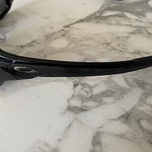 May include: Black sunglasses with a glossy finish. The sunglasses have a curved design with a dark lens and a sleek, modern aesthetic. The frame features a subtle logo detail.