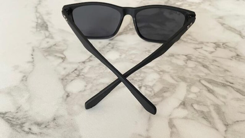 Pu&ograve; includere: Occhiali da sole neri con lenti scure. Le aste degli occhiali da sole sono incrociate, appoggiate su una superficie di marmo bianco e grigio. Gli occhiali hanno una montatura rettangolare.