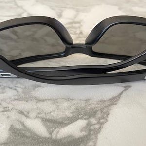 May include: Black sunglasses with a sleek, modern design. The frames are a glossy black, and the lenses are dark gray. The sunglasses are resting on a marble-patterned surface.