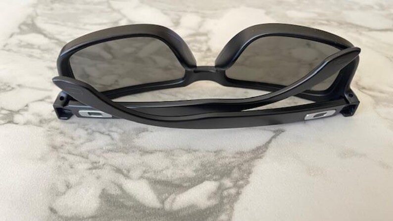 May include: Black sunglasses with a sleek, modern design. The frames are a glossy black, and the lenses are dark gray. The sunglasses are resting on a marble-patterned surface.