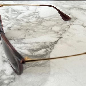 May include: Sunglasses with a dark burgundy frame and tinted lenses. The temples are a thin gold color, transitioning to a matching burgundy at the ear pieces. The glasses are set against a marble background.