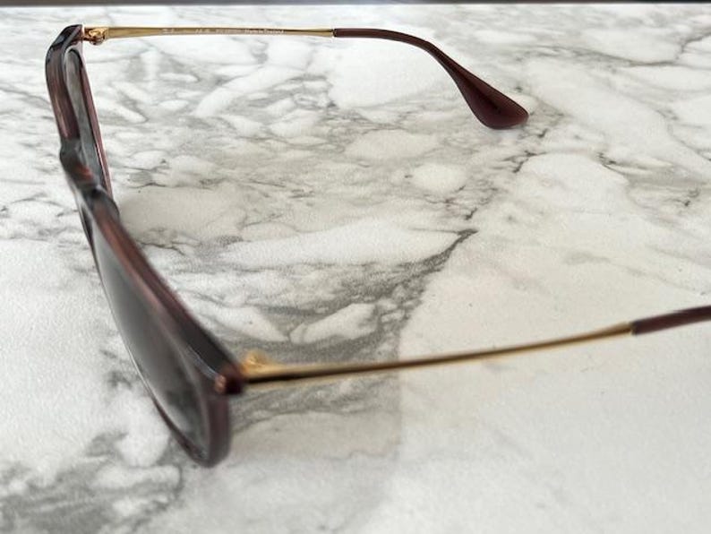 May include: Sunglasses with a dark burgundy frame and tinted lenses. The temples are a thin gold color, transitioning to a matching burgundy at the ear pieces. The glasses are set against a marble background.
