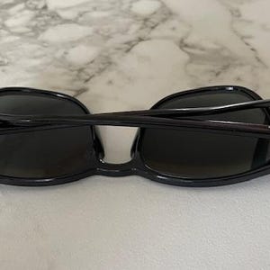 Puede incluir: Gafas de sol negras con lentes oscuras, vistas desde arriba. Las monturas son de color negro brillante y las patillas est&aacute;n plegadas. Las lentes son ovaladas y las gafas est&aacute;n sobre una superficie clara.