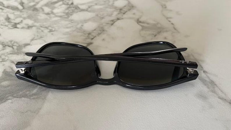 Puede incluir: Gafas de sol negras con lentes oscuras, vistas desde arriba. Las monturas son de color negro brillante y las patillas est&aacute;n plegadas. Las lentes son ovaladas y las gafas est&aacute;n sobre una superficie clara.