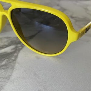 May include: Bright yellow sunglasses with dark gradient lenses. The sunglasses have a thick frame and a classic aviator shape. The Ray-Ban logo is visible on the temple.