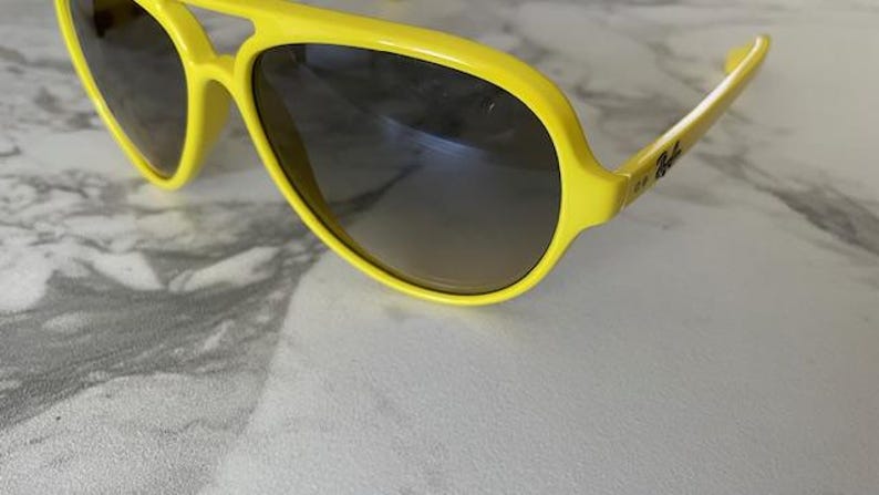 May include: Bright yellow sunglasses with dark gradient lenses. The sunglasses have a thick frame and a classic aviator shape. The Ray-Ban logo is visible on the temple.