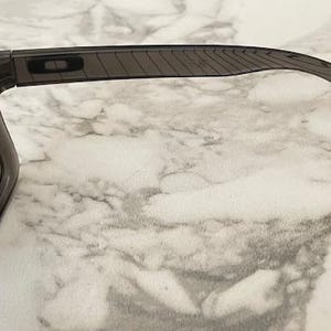May include: Dark gray sunglasses with a rectangular frame and tinted lenses. The temples are a lighter gray with a textured design and a small red accent at the end. The sunglasses are set against a marble background.